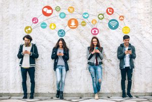 Social network concept with smiling young people holding smart phones in front of big white wall.